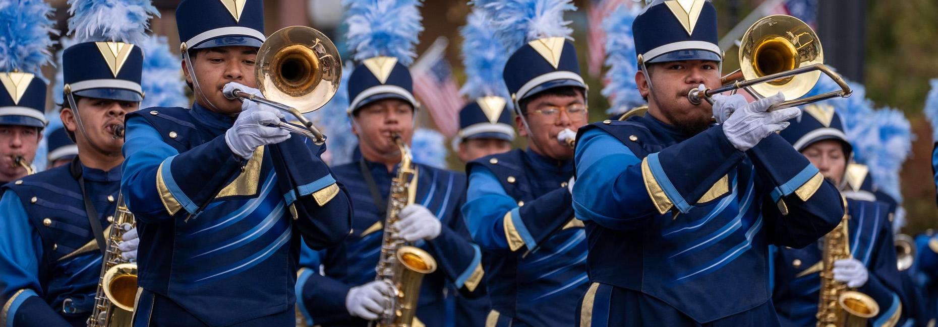 Members of a school marching band perform during a parade in blue uniforms with feathered plumes.