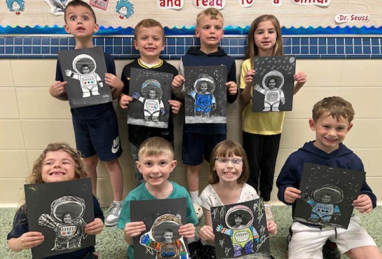 8 students hold artwork they created which features their faces on an astronaut's body