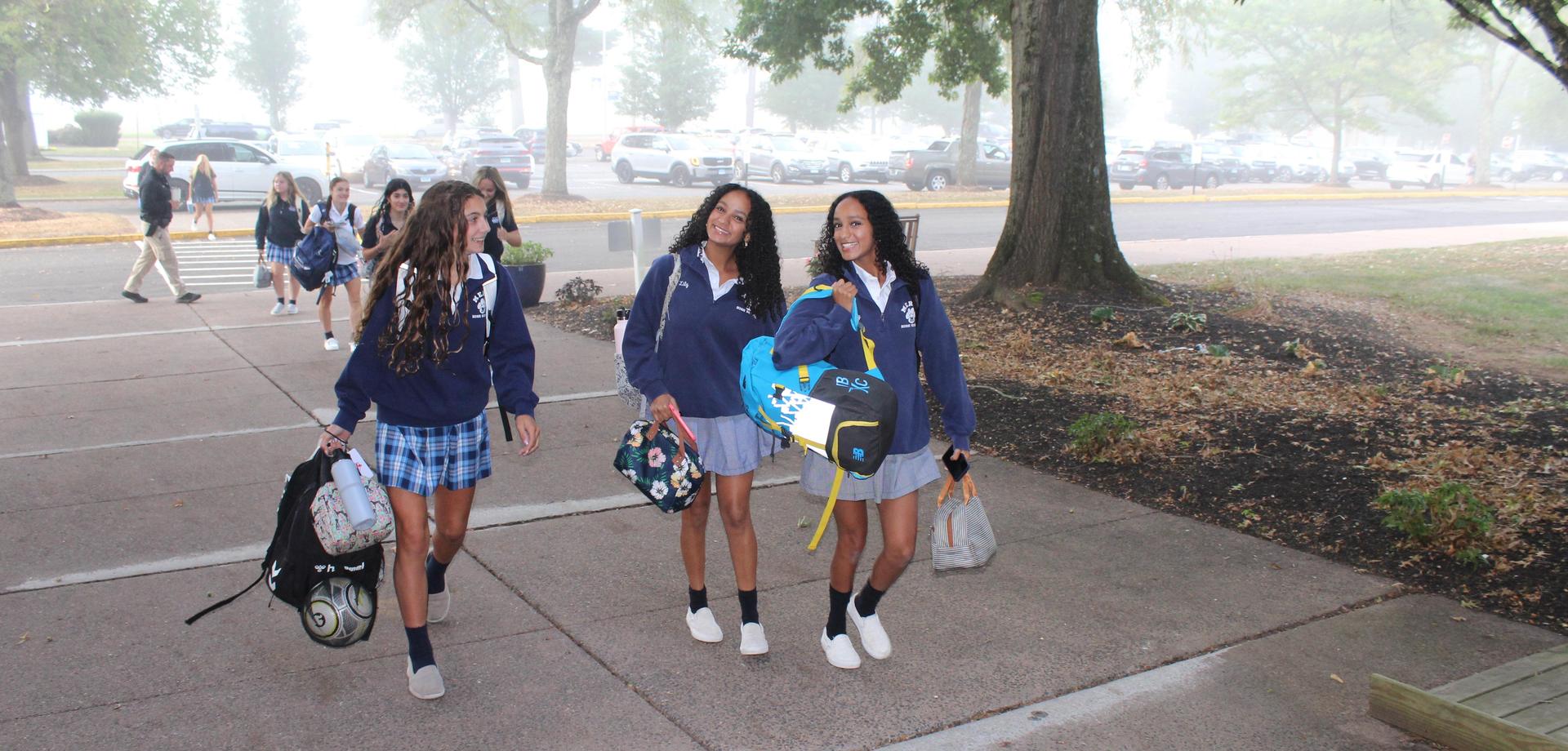 Three Mercy students in school uniforms smiling and walking together into school on the first day of classes.
