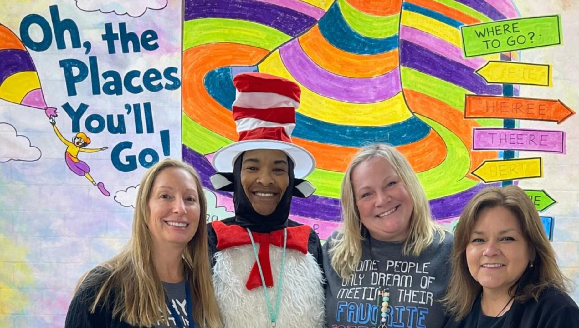 Four women pose together, one dressed as a penguin, in a colorful classroom.