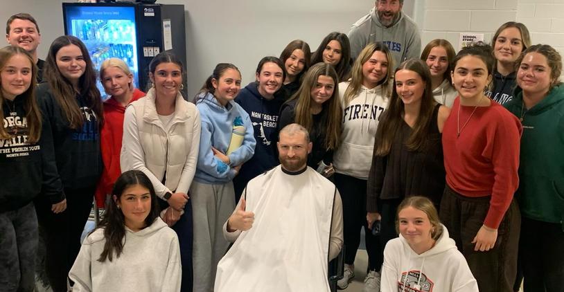 A group of smiling girls posing with a man in a barber's cape giving a thumbs up.