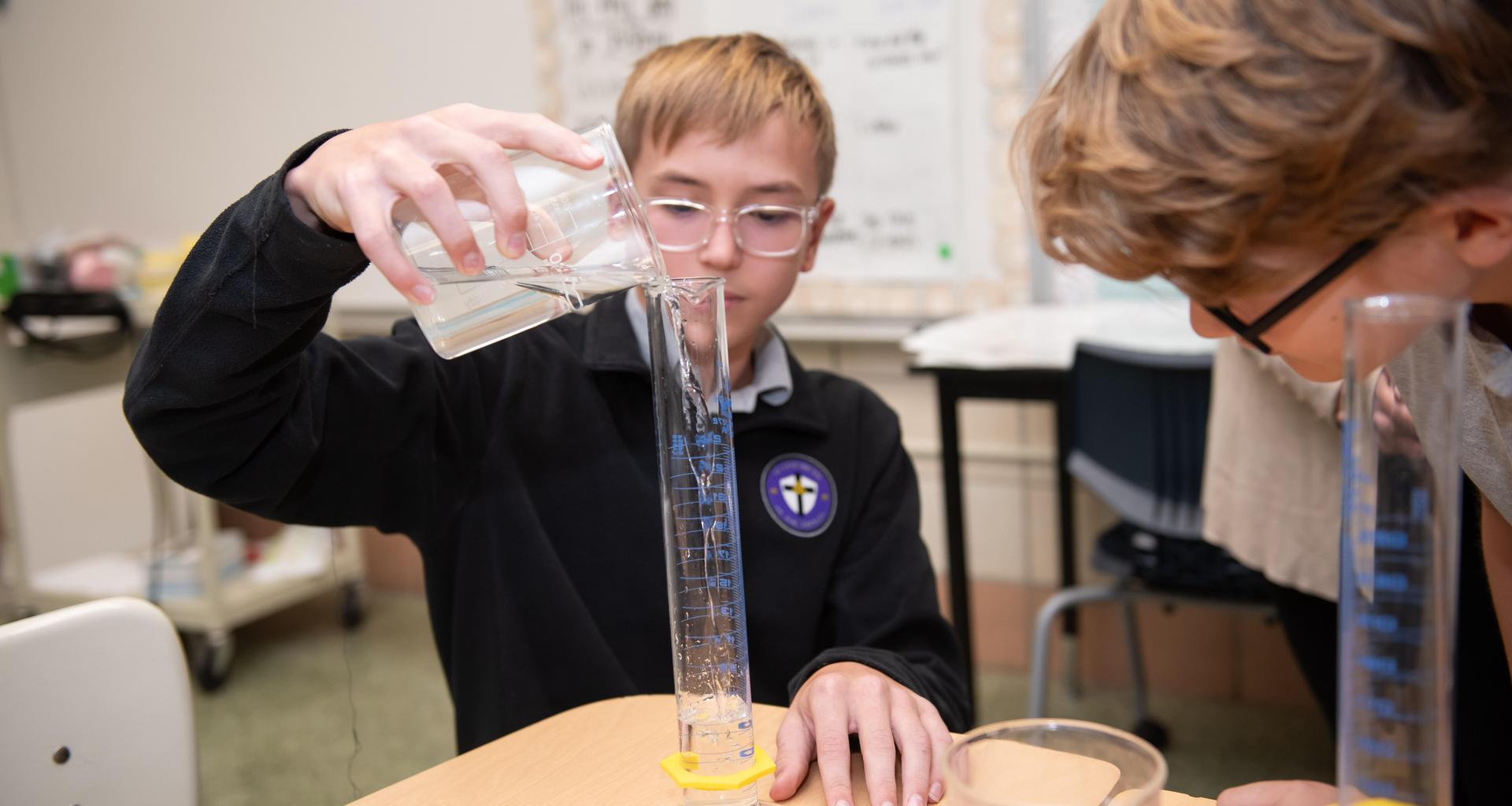 Child pouring liquid into a measuring cylinder, another observing closely.