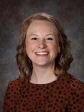Smiling teacher with wavy hair wearing a black polo shirt against a gray backdrop.