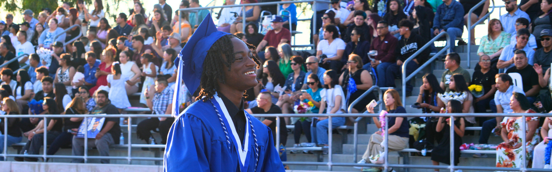 A joyful graduate in a blue cap and gown smiles at a crowded graduation ceremony.