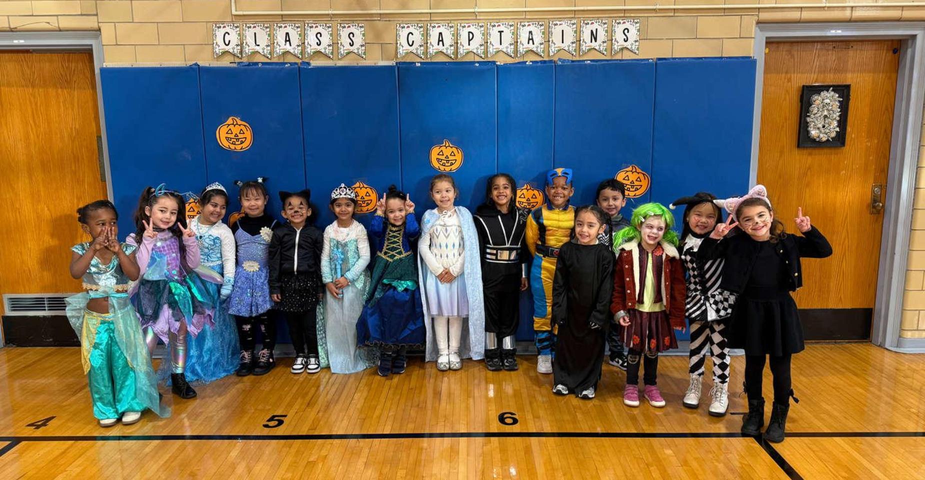 Children in costumes pose together in a school gym, smiling for a group photo.