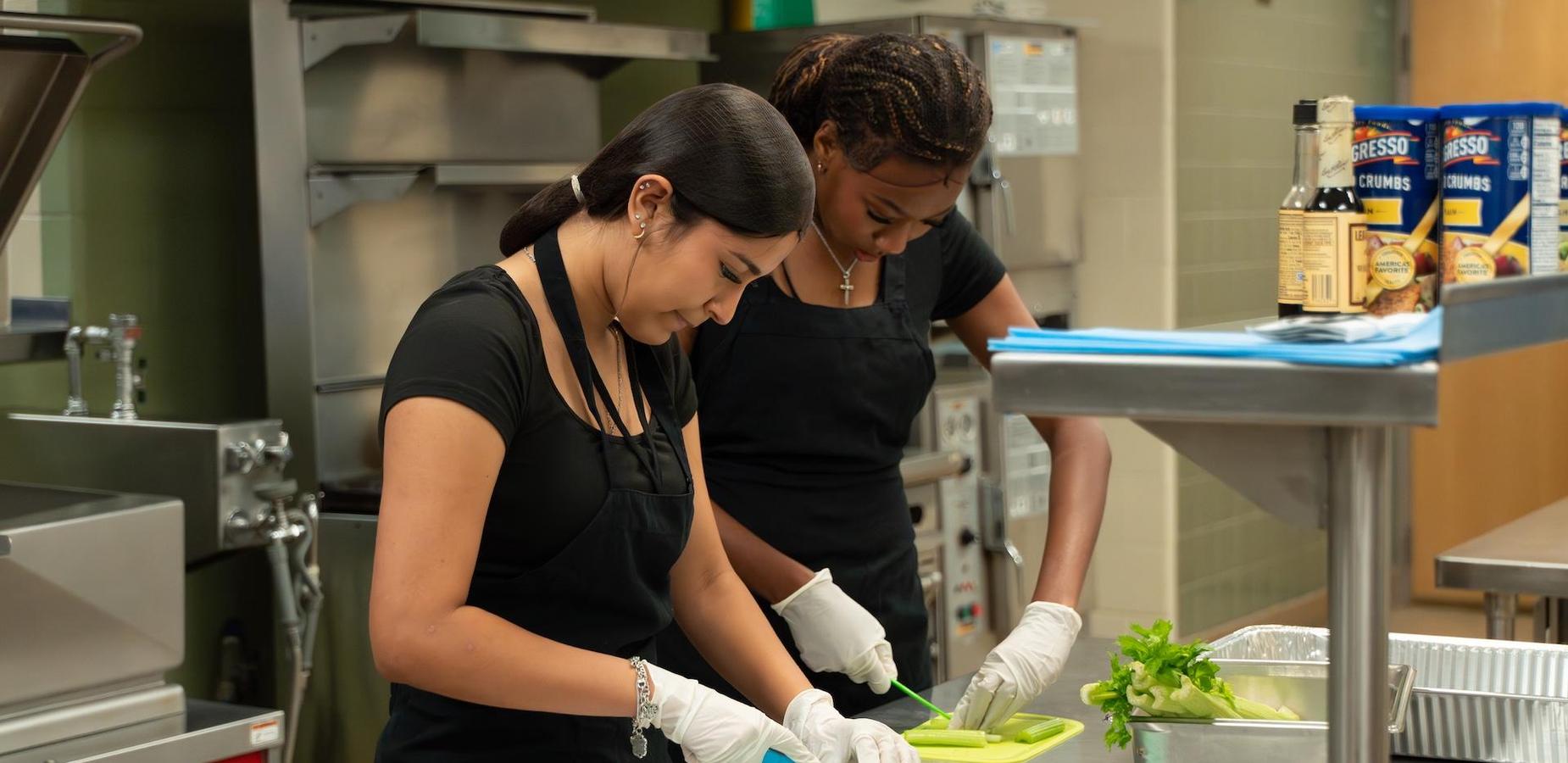 Two women in black aprons preparing food in a kitchen environment.