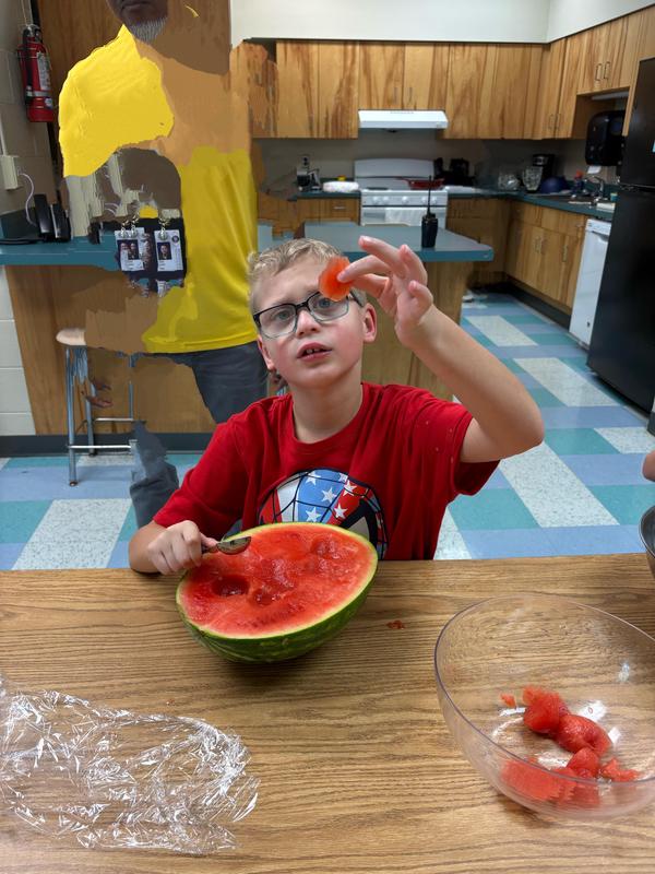 A student in a red shirt looks closely at a piece of watermelon while working on his fruit bowl.