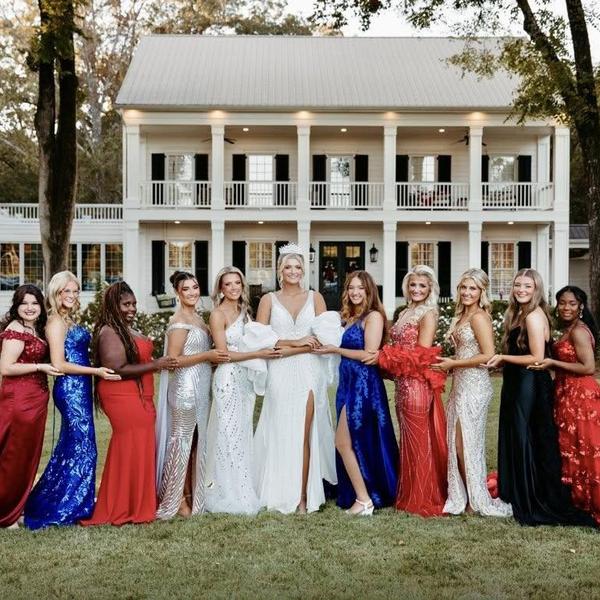 A group of women in elegant dresses posing in front of a large white house.