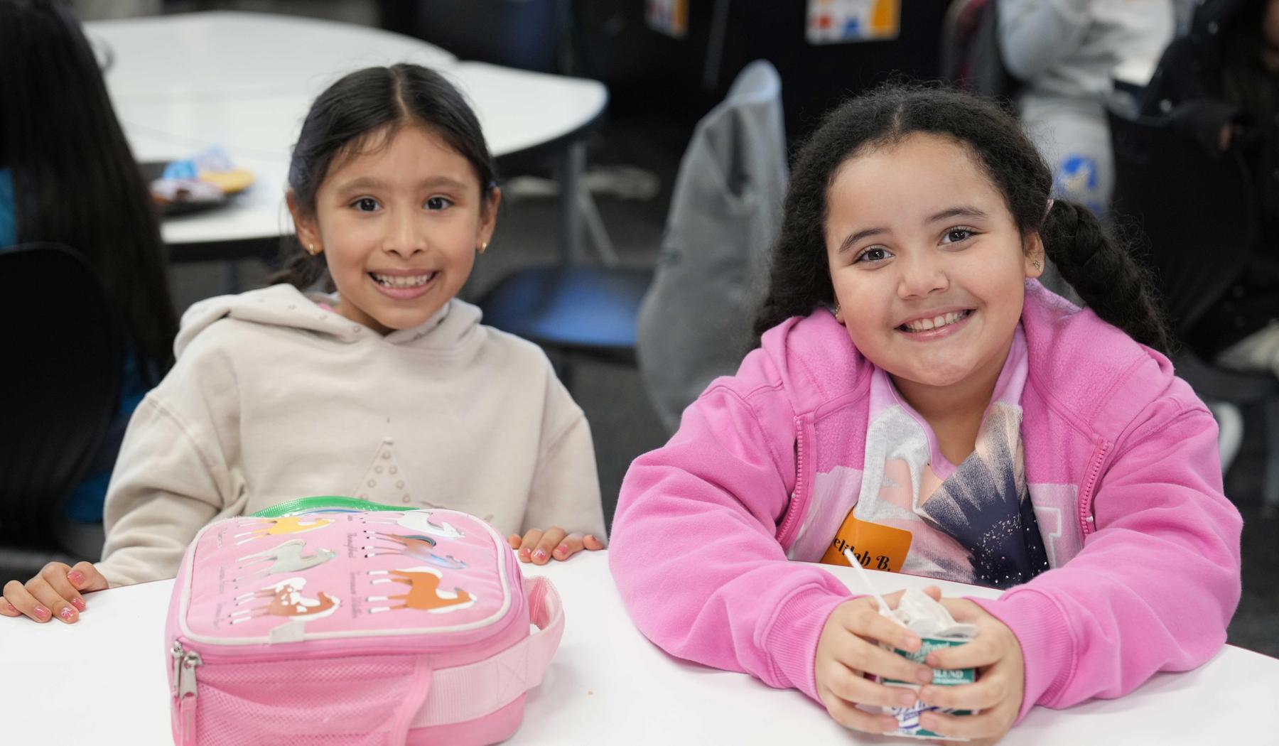 two smiling girls sitting. One with lunch bag and a the other with a carton of milk