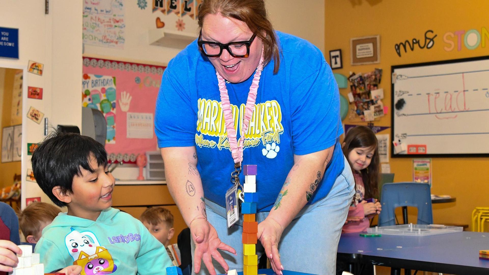 Teacher helps a boy build a tower with blocks in a colorful classroom.