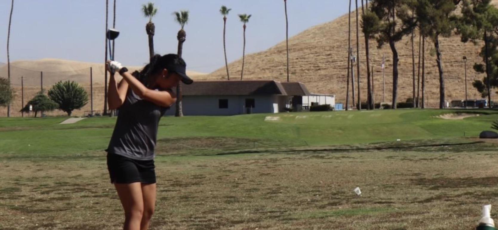 A golfer swings at the driving range with palm trees and hills in the background.