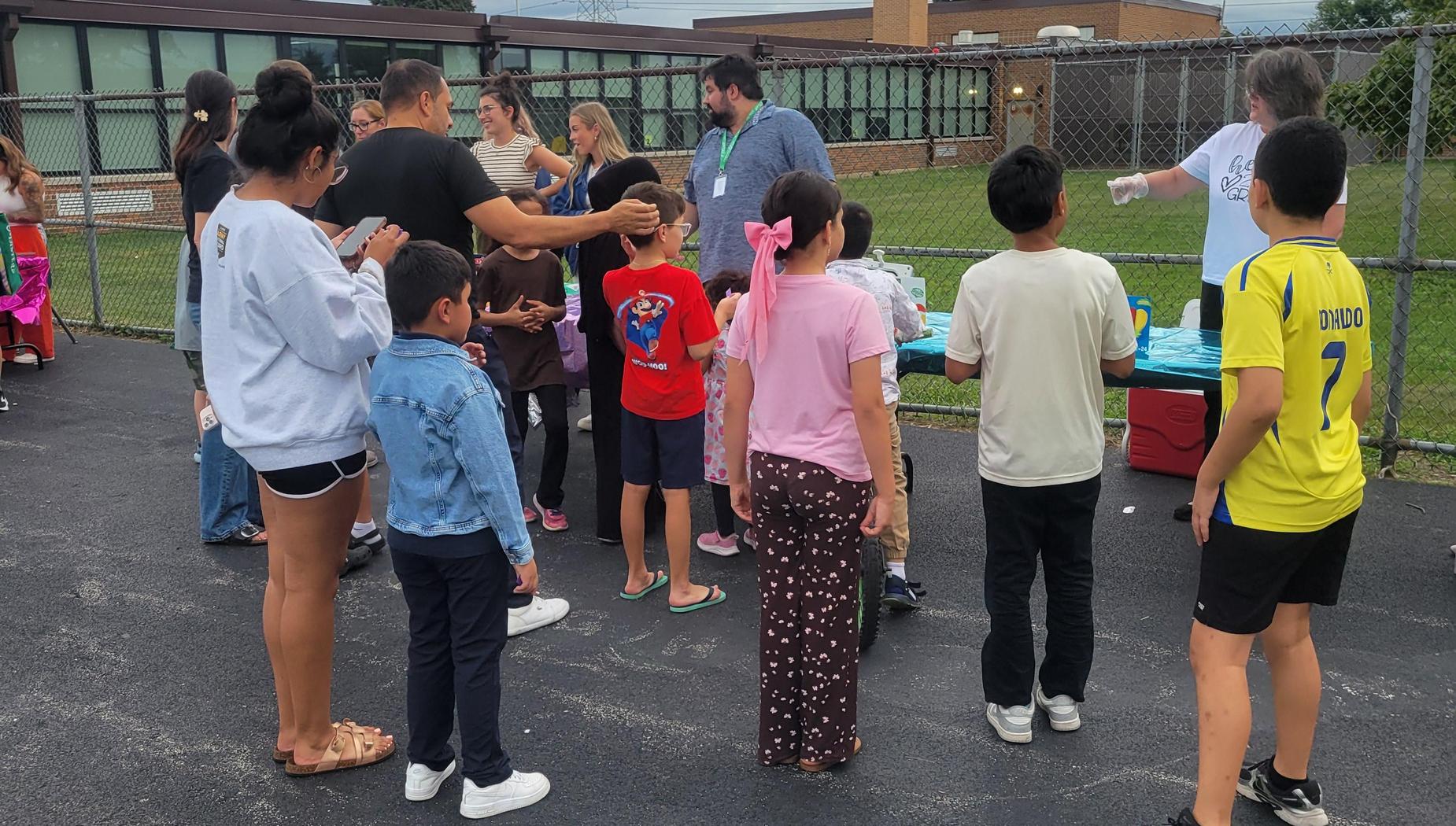 A group of children and adults waiting in line outside by a school for refreshments.