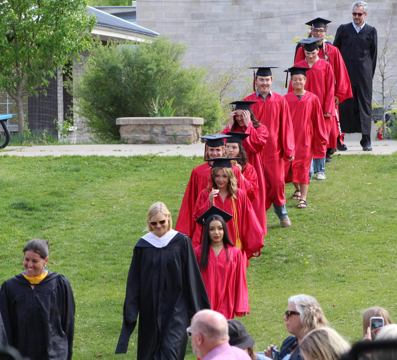Graduating students in red gowns and caps walking on a grassy hill with an educator.