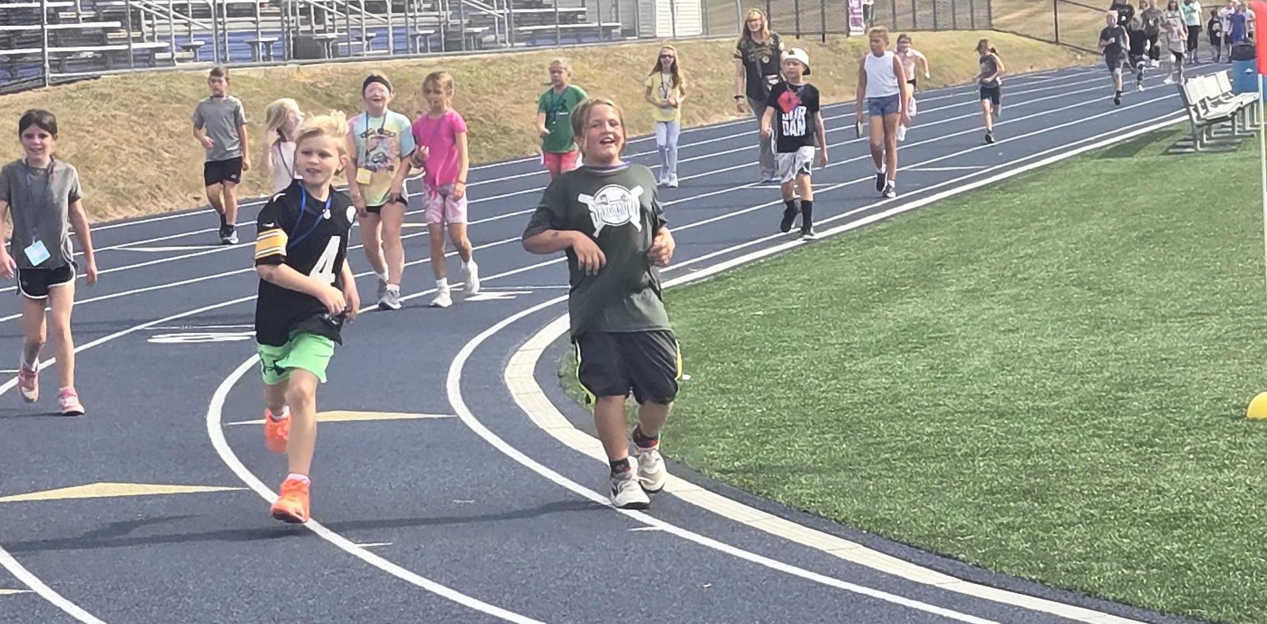 Two children running joyfully on a track surrounded by others.