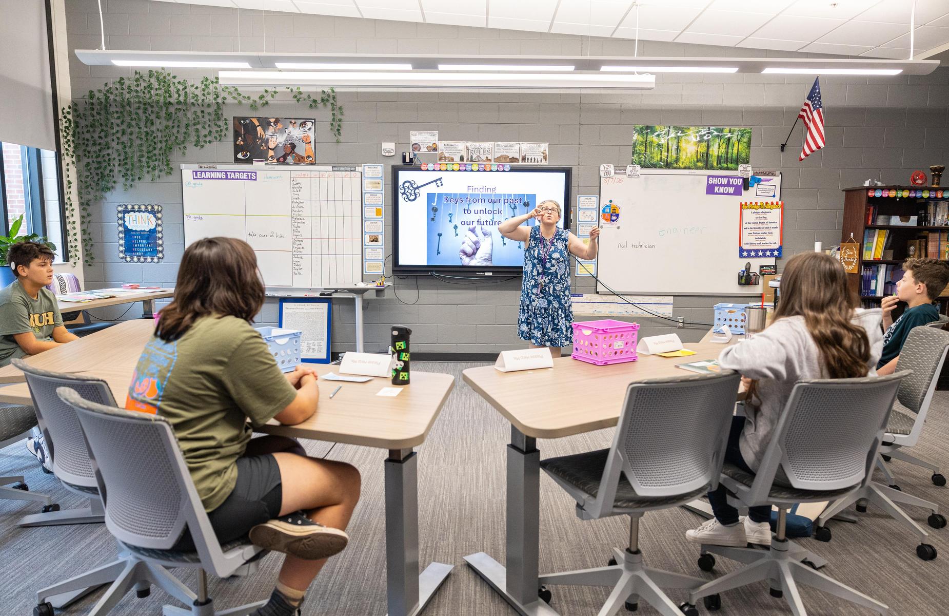 A teacher presenting to students seated around a classroom table with a screen display.