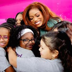 A group of girls embracing a smiling woman against a vibrant pink background.