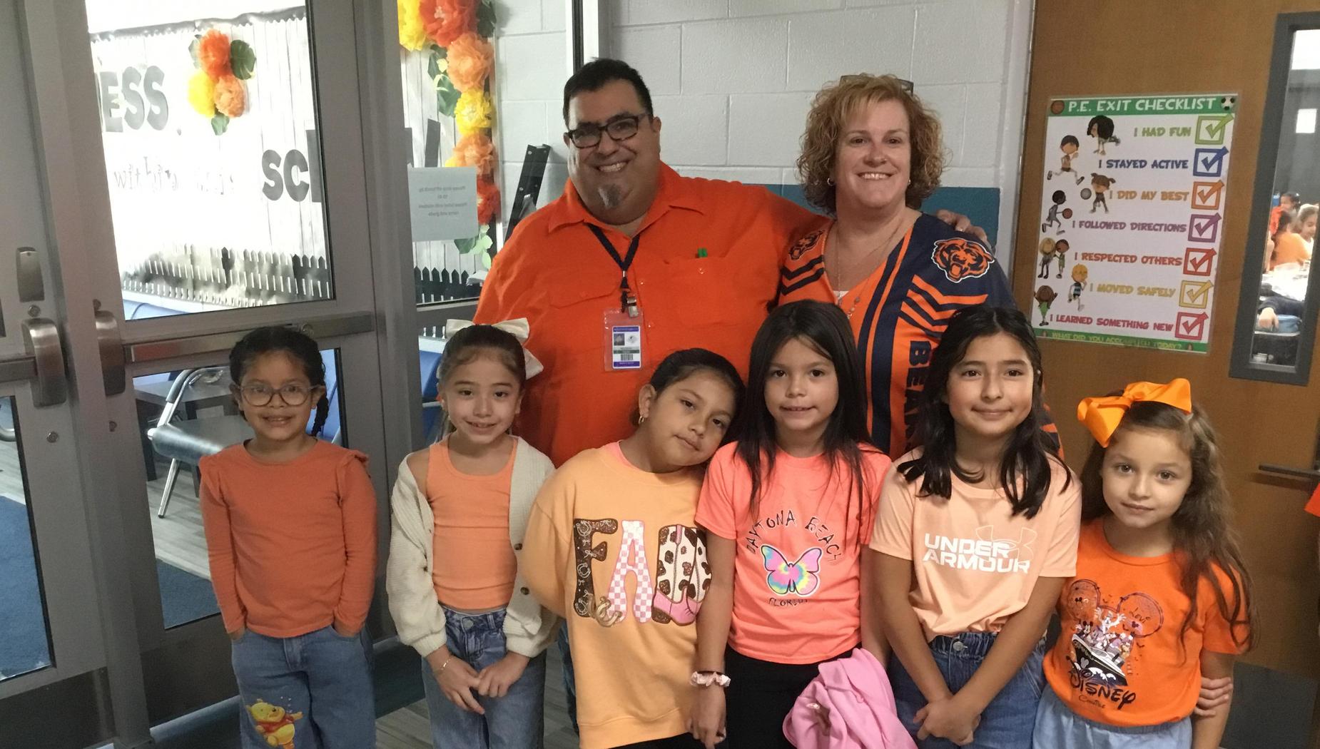 Group of children and adults wearing orange clothes, smiling at a school entrance.