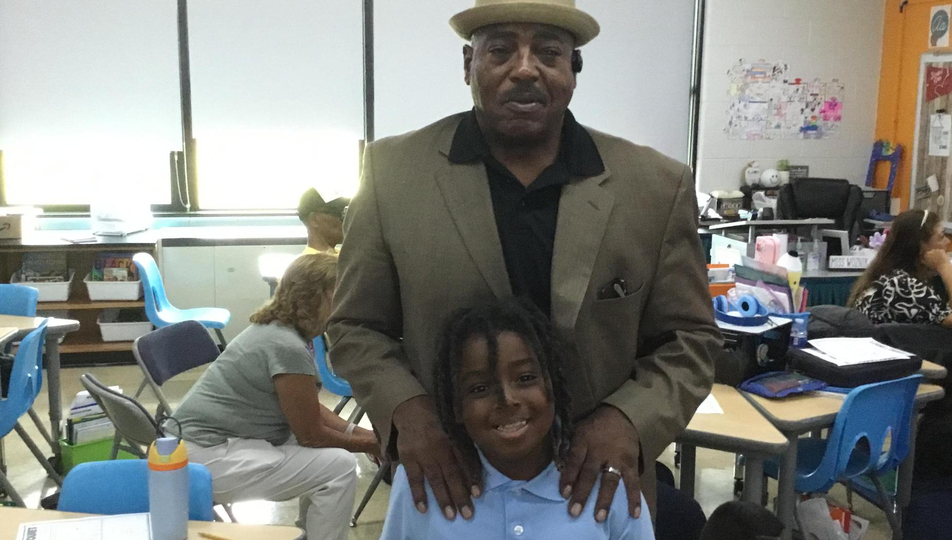 A man in a hat standing with a young boy in a classroom with peers in the background.