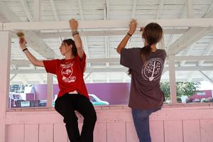 Two students sit on a railing painting a white overhead beam inside of a livestock pen