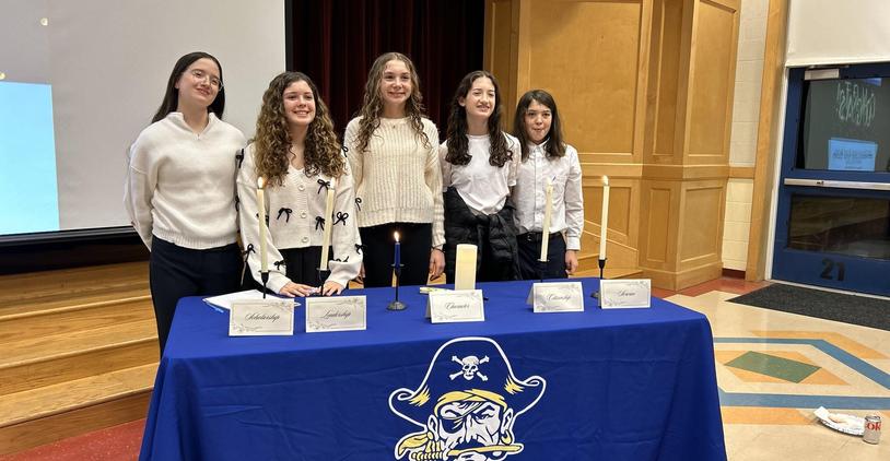 Five girls in white sweaters standing behind a table with candles and a blue pirate-themed tablecloth.
