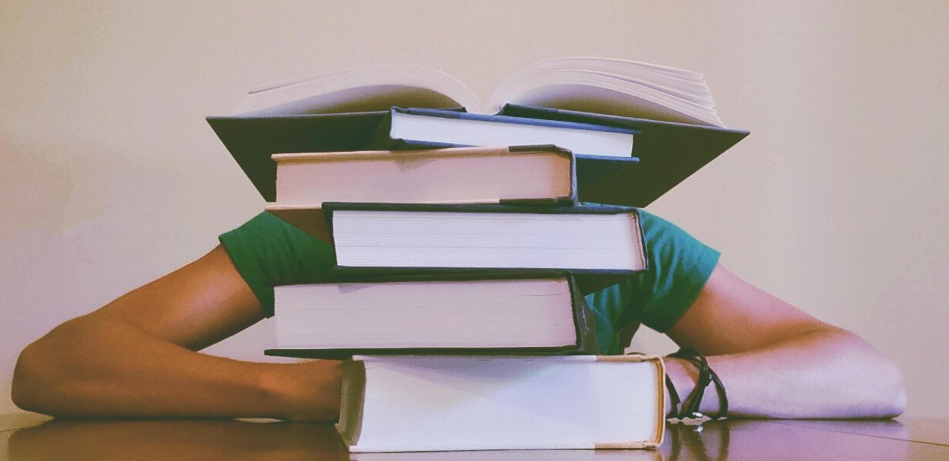 A person sitting at a table with a stack of books obscuring their face.