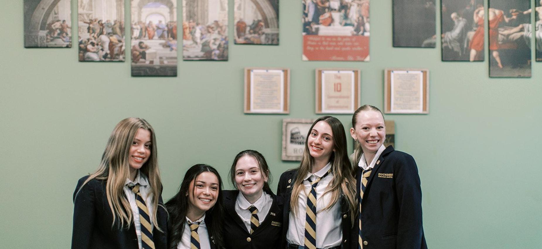Five smiling high school students in blazers stand together in front of framed art
