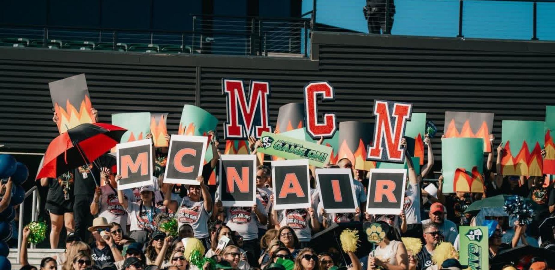 A crowd holds up large signs spelling 'MCNAIR', surrounded by colorful decorations.