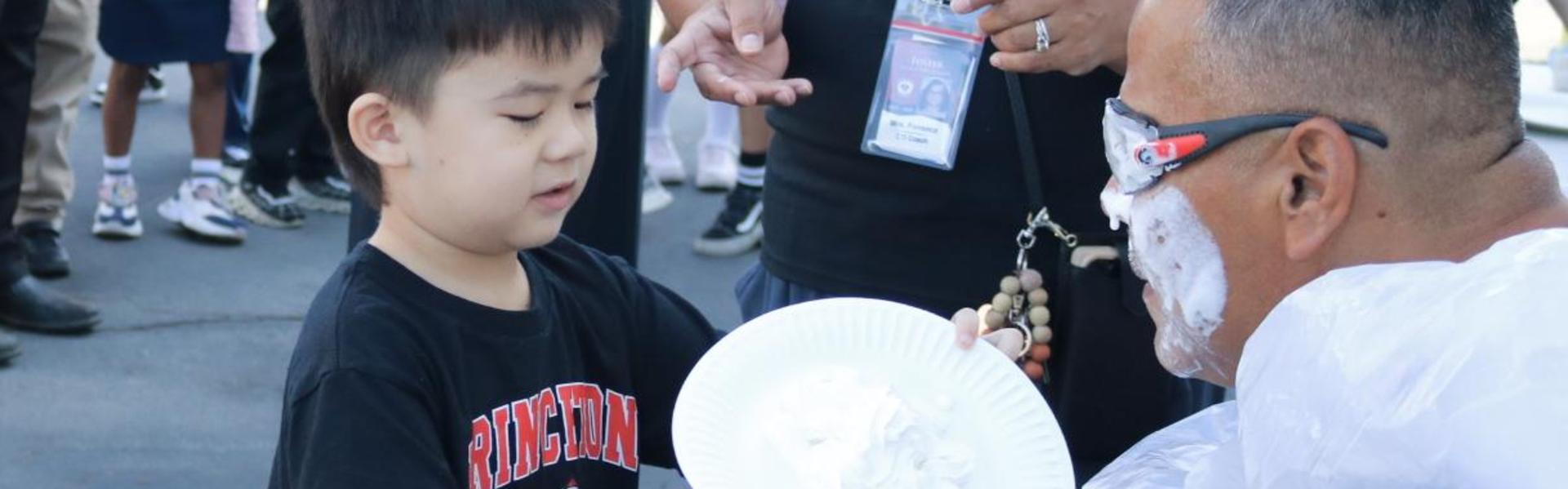 A child hands a plate to an adult covered in whipped cream at a school event.