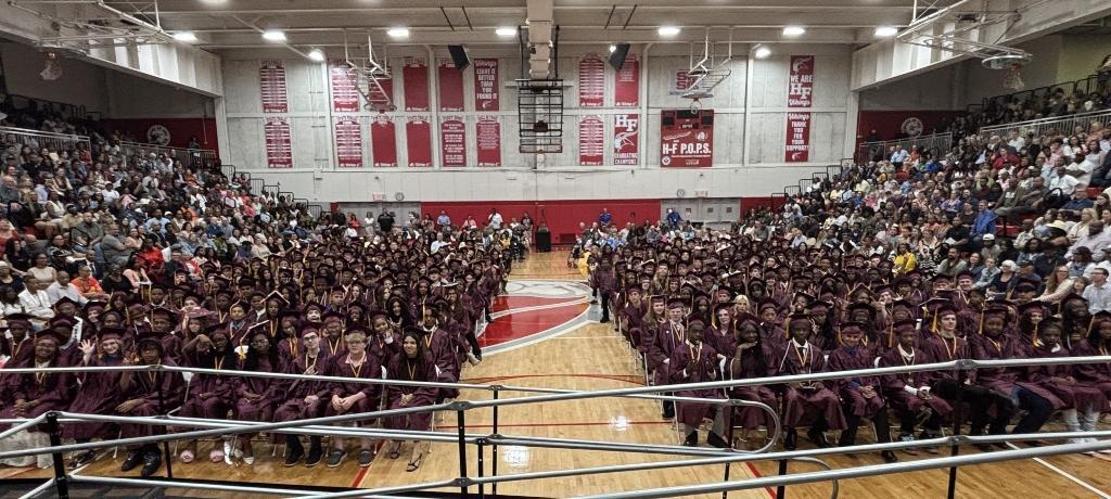 gym filled with graduates in caps and gowns and loved ones in the bleachers