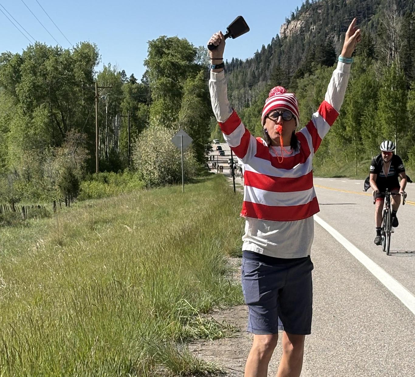 A person in a striped shirt and hat celebrates on the roadside during a cycling event.