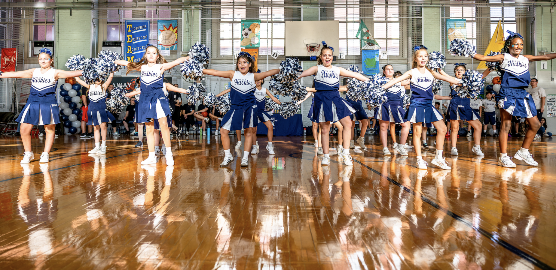 Cheerleaders performing in a gymnasium with wooden floors and banners.