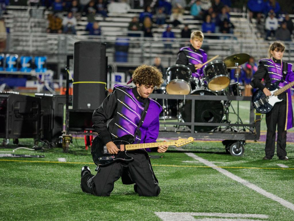 A Marching Band member on his knees playing guitar on the football field during a performance.