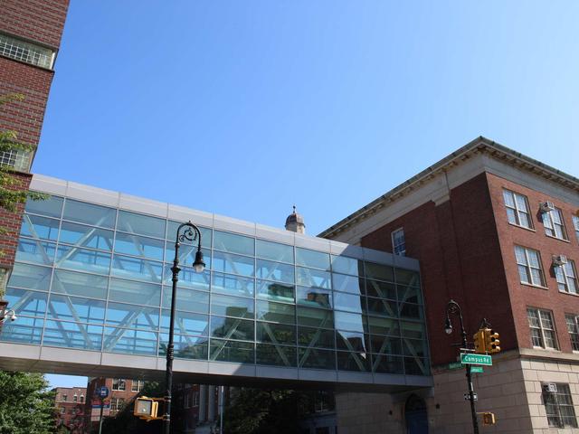 Modern glass skybridge connecting two brick buildings under a clear blue sky.