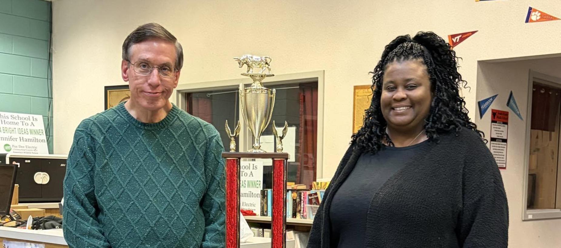 Two individuals standing beside a large trophy in a library setting.