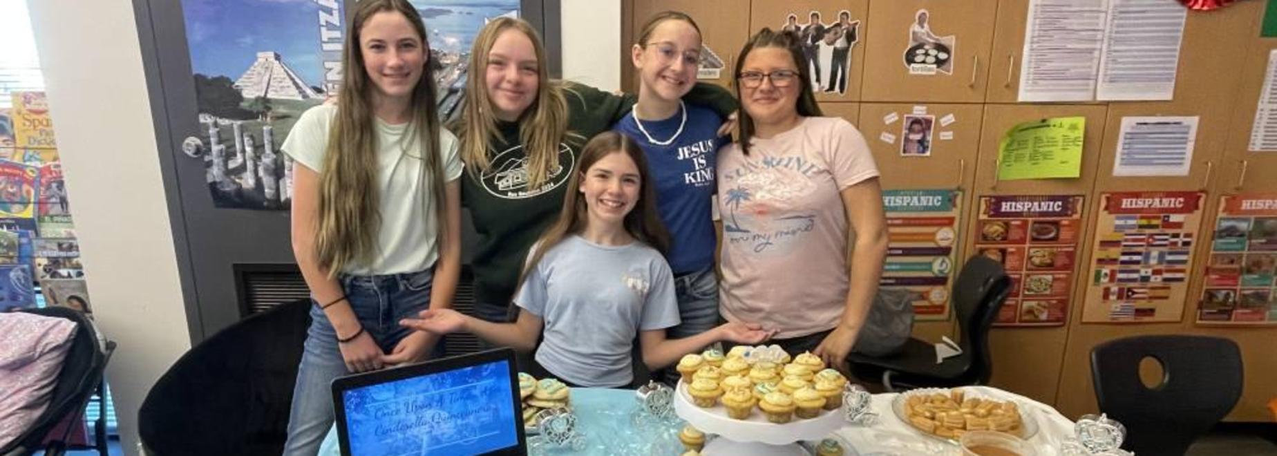 Five girls posing beside a table with cupcakes and a laptop during an event.
