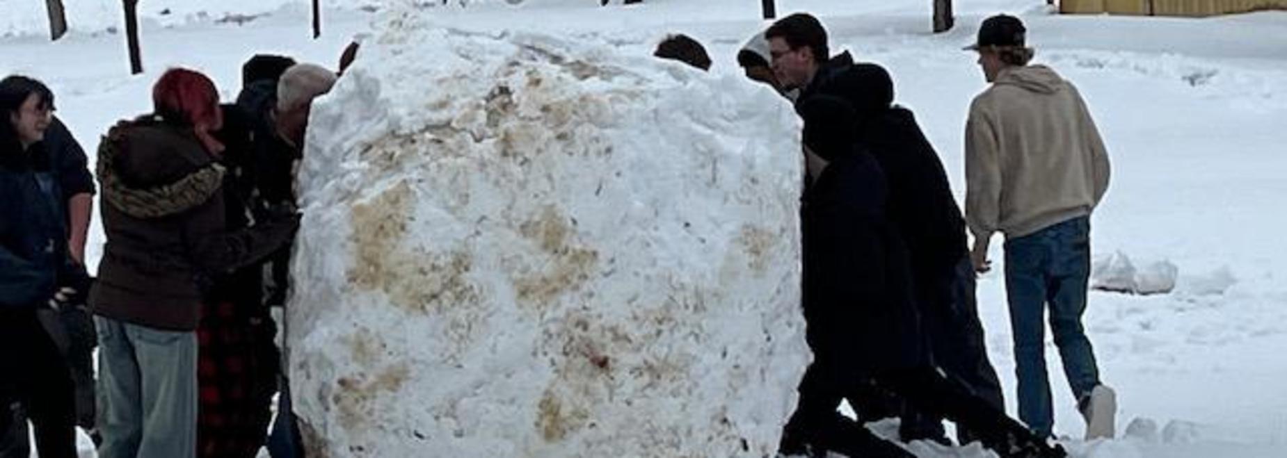 Group of people pushing a large snowball in a snowy landscape.