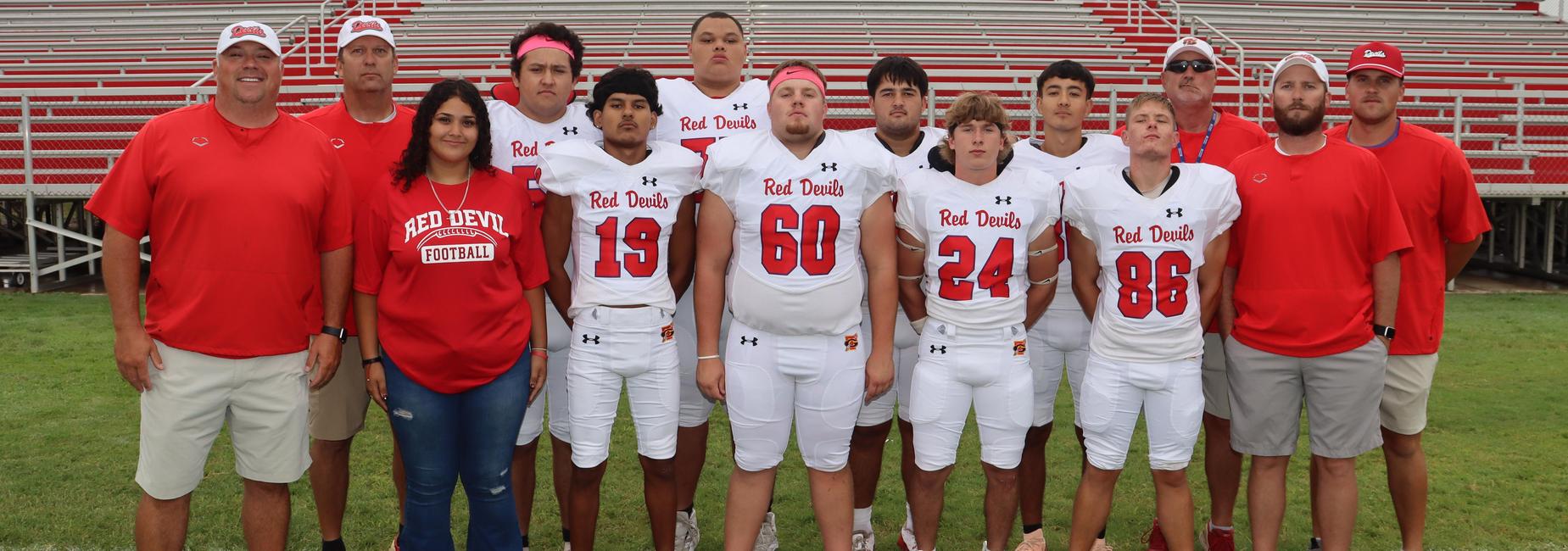 A football team with coaches, wearing red and white uniforms, posing in front of bleachers.