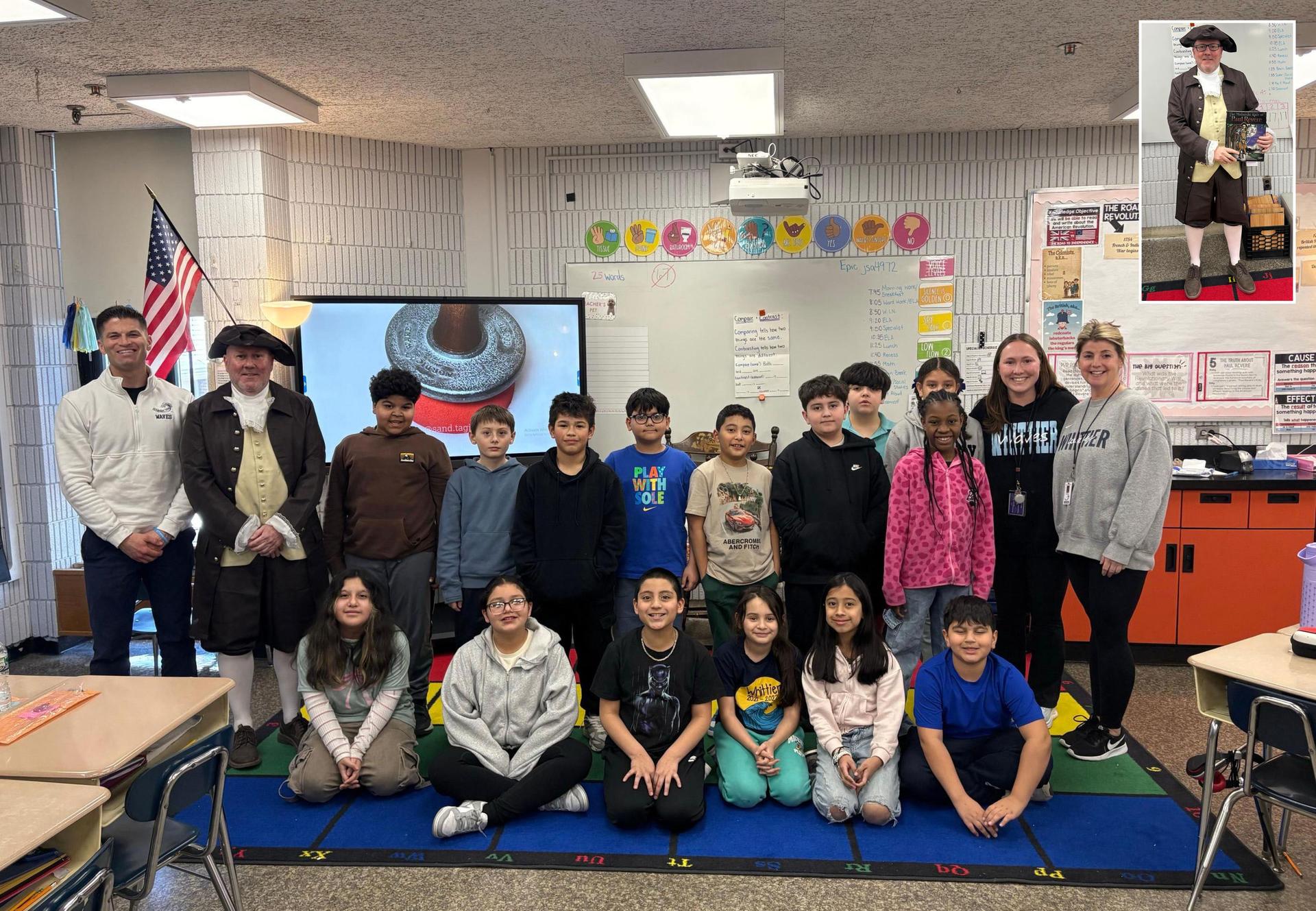A class of students poses with a teacher and a guest in historical attire in a classroom.