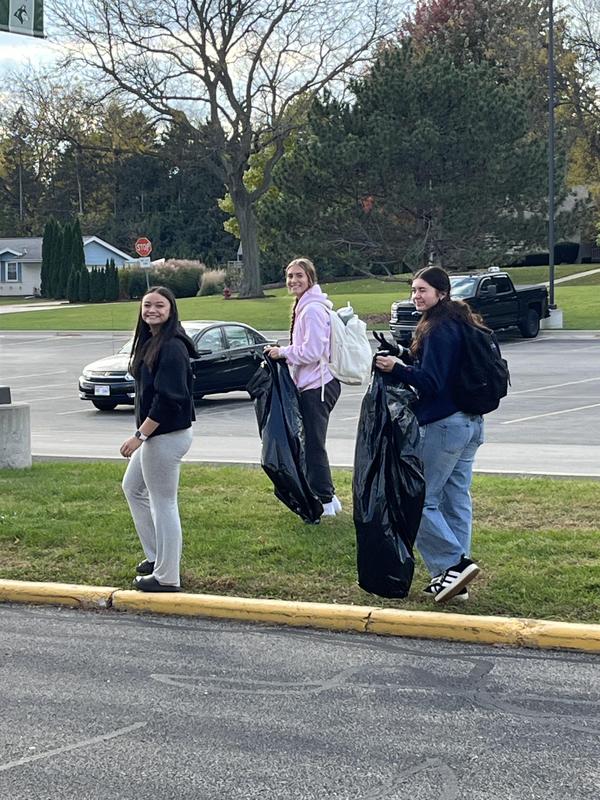 Three young women walking with bags of trash down a grassy area by the road.