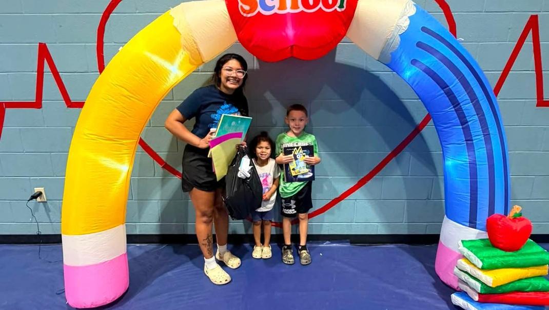 A smiling woman and two children stand under a colorful school-themed arch.