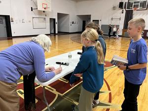 A woman signs a book for children at a table in a gym, with kids watching eagerly.