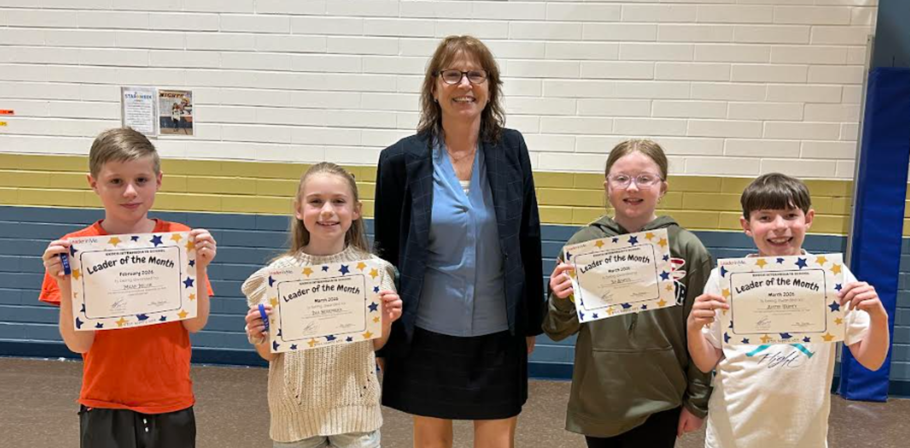 Four students holding 'Leader of the Month' certificates with an adult in a gym.
