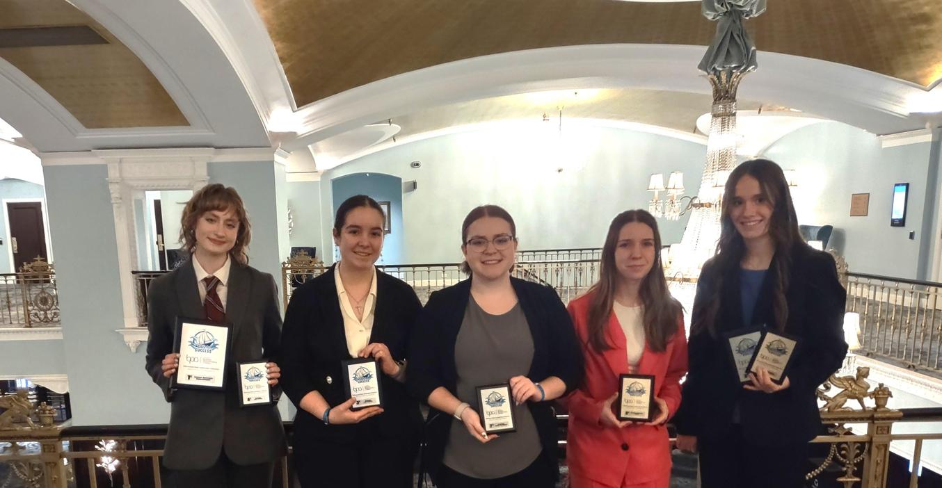 Five women in business attire holding awards, standing in a hotel lobby.