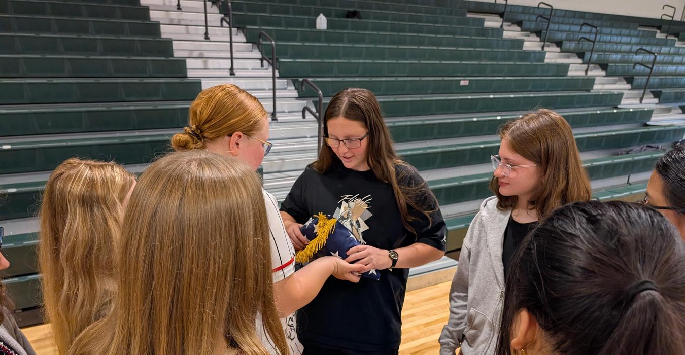 A group of girls examines a flag while engaged in a discussion in a gym.