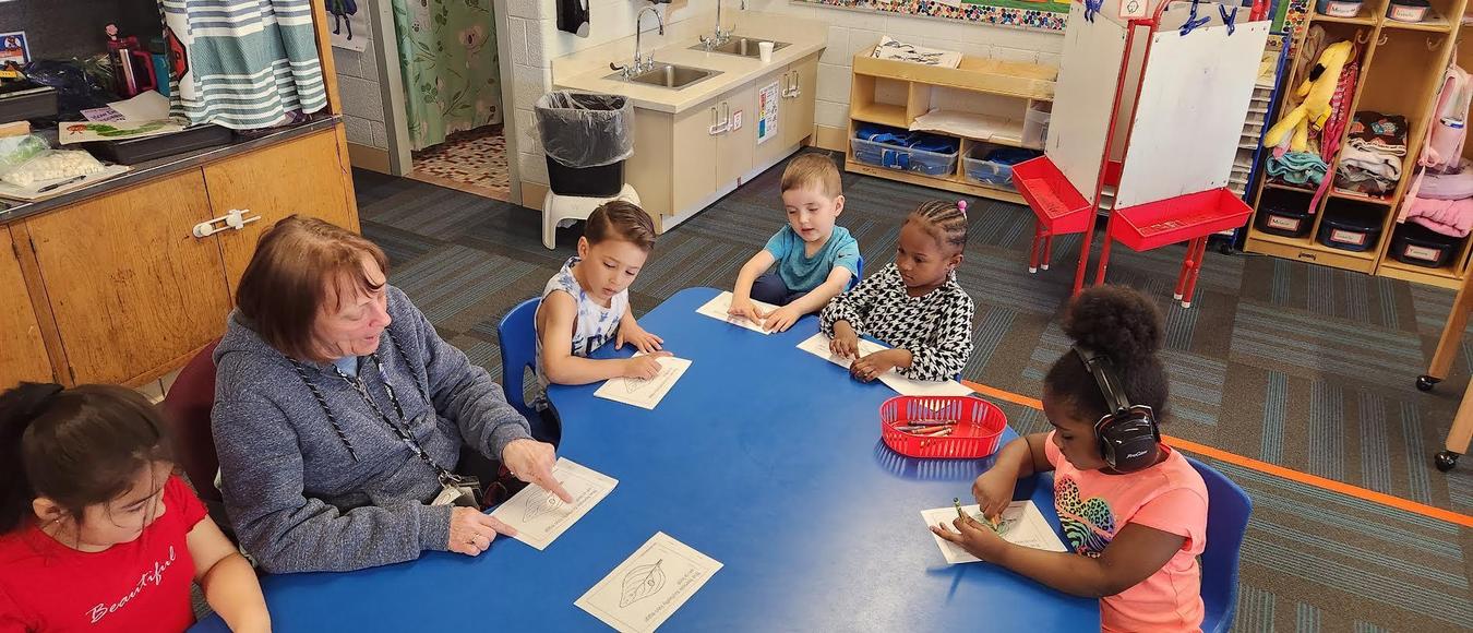 Children are sitting at a blue table with a teacher practicing their reading skills.