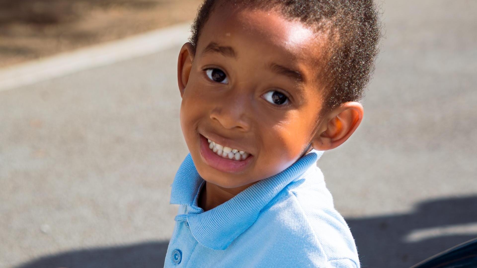 A boy in a light blue shirt grins at the camera, sitting outdoors.