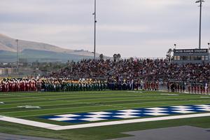 Multiple school marching bands in colorful uniforms lined up on a football field performing for a packed crowd.