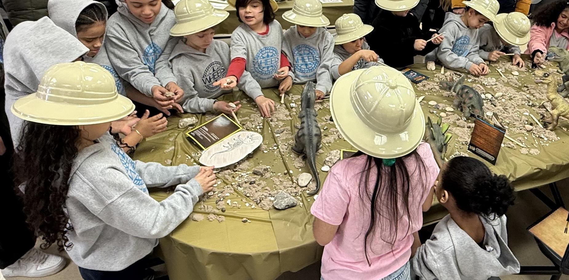 Children in hats explore a fossil dig with dinosaur models and rocks on a table.