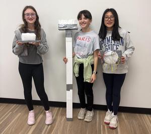 three students standing next to a styrofoam broom.