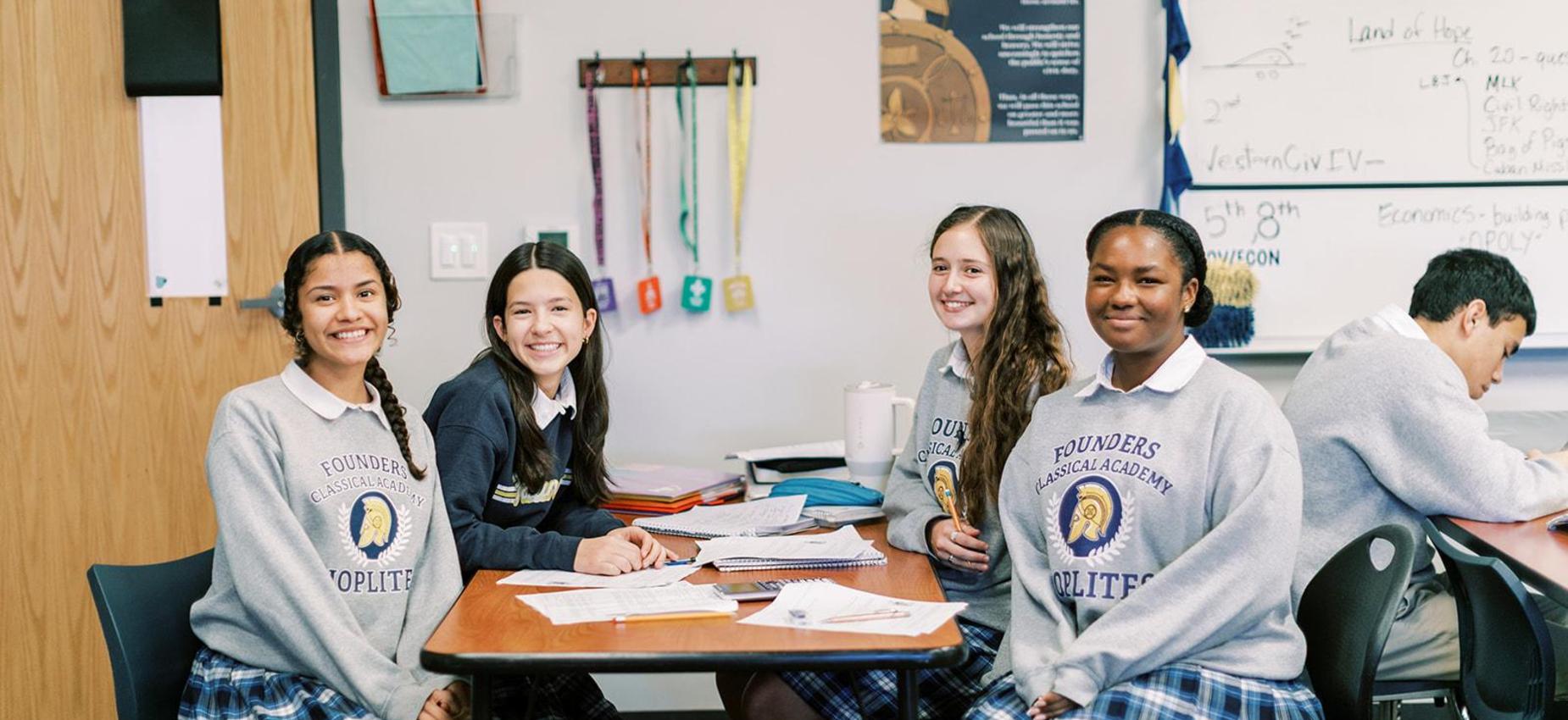Smiling high school students in plaid uniform skirts sit together at a classroom table